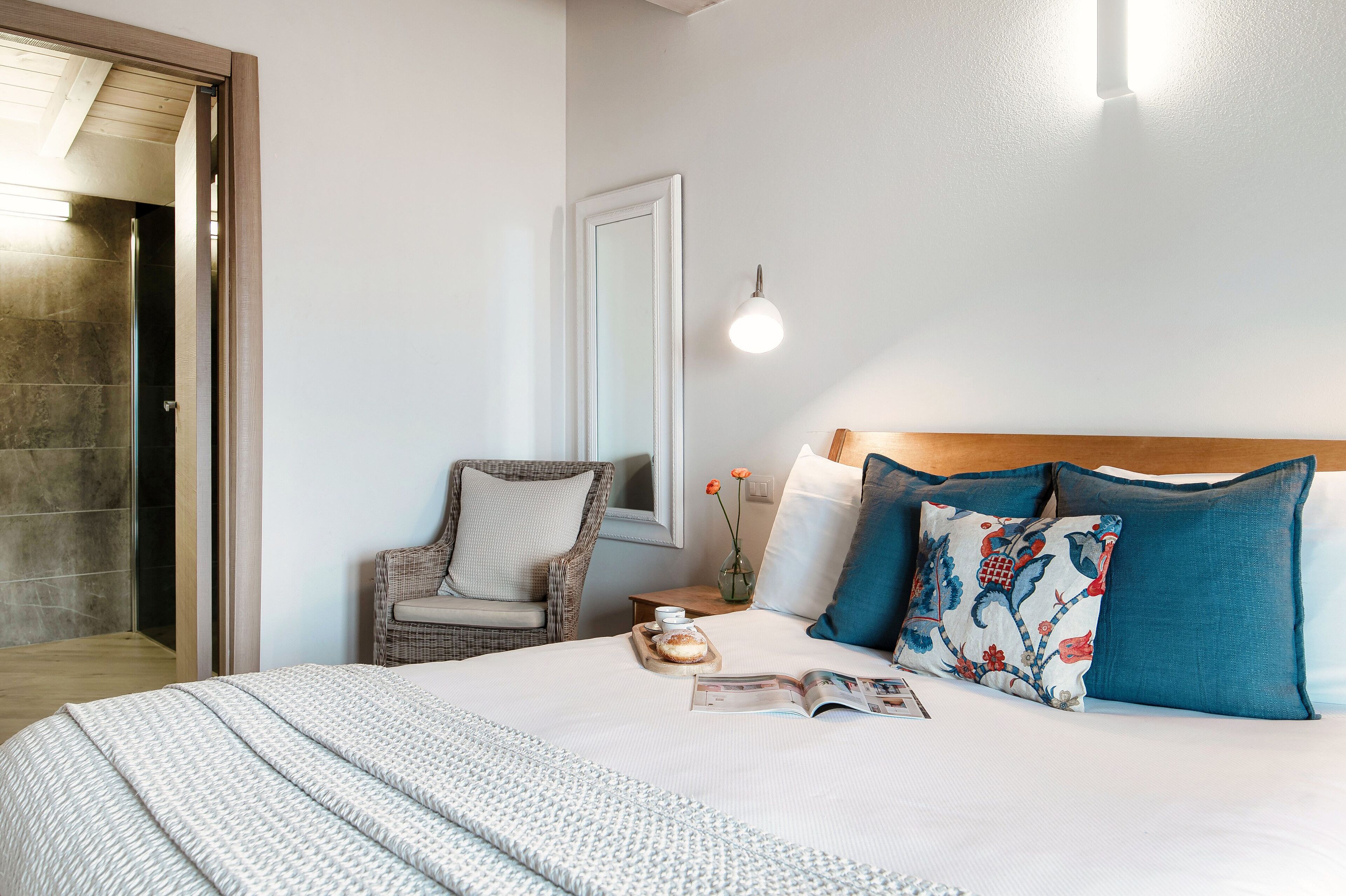 Cama bem arrumada com edredom branco, bandeja com xícara e pires, cadeira de vime com almofada, mesa de cabeceira com um vaso de flores e luminária fixada na parede.