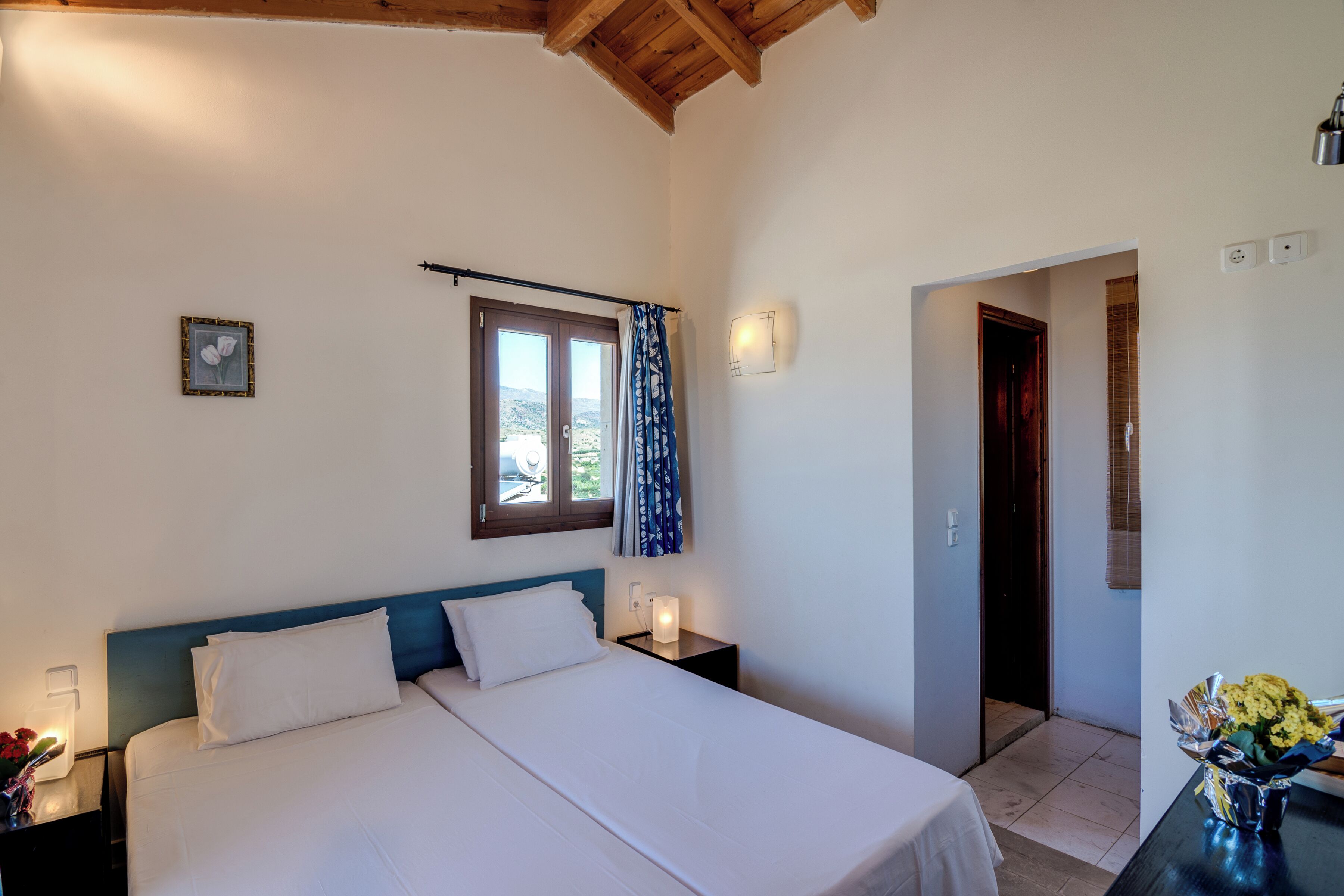 Un dormitorio con cama, mesitas de noche, una ventana con cortinas, una lámpara de pared y una puerta que da a otra habitación.