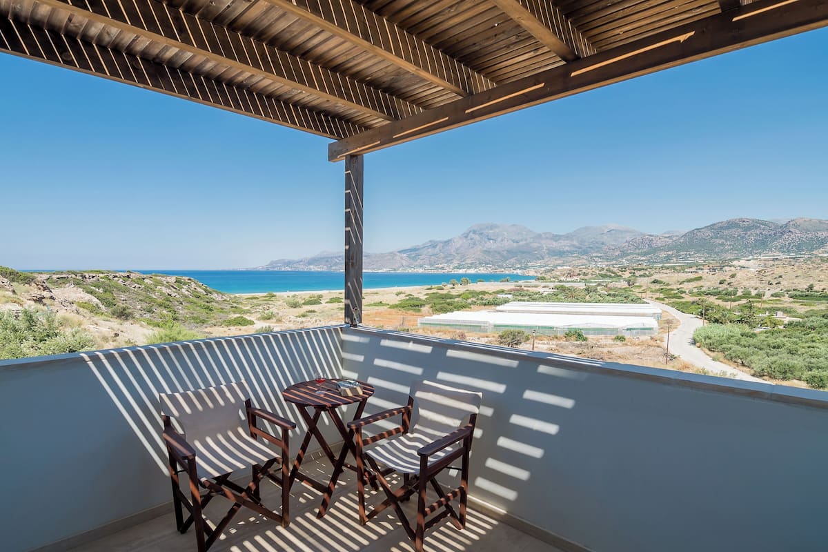 A balcony with a table and chairs, overlooking a coastal landscape with mountains and a clear blue sky.
