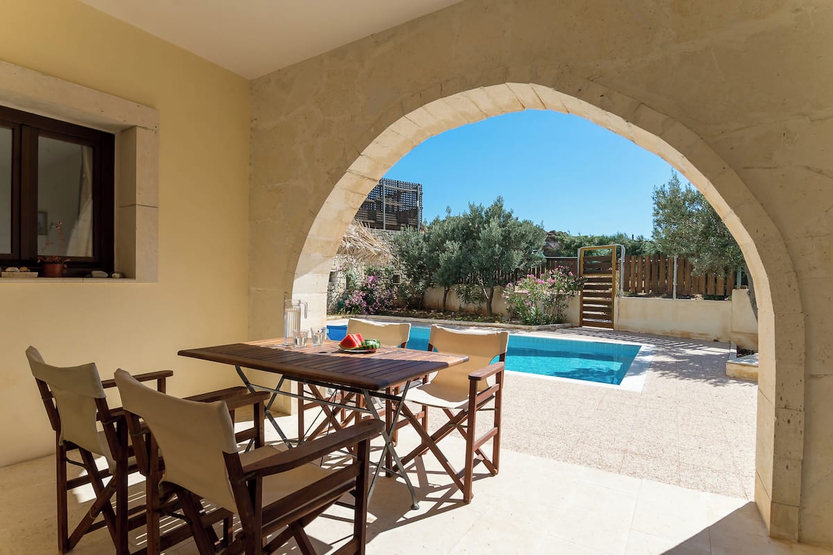 An outdoor patio with a wooden table and chairs, a swimming pool, and a view of the surrounding garden and trees.