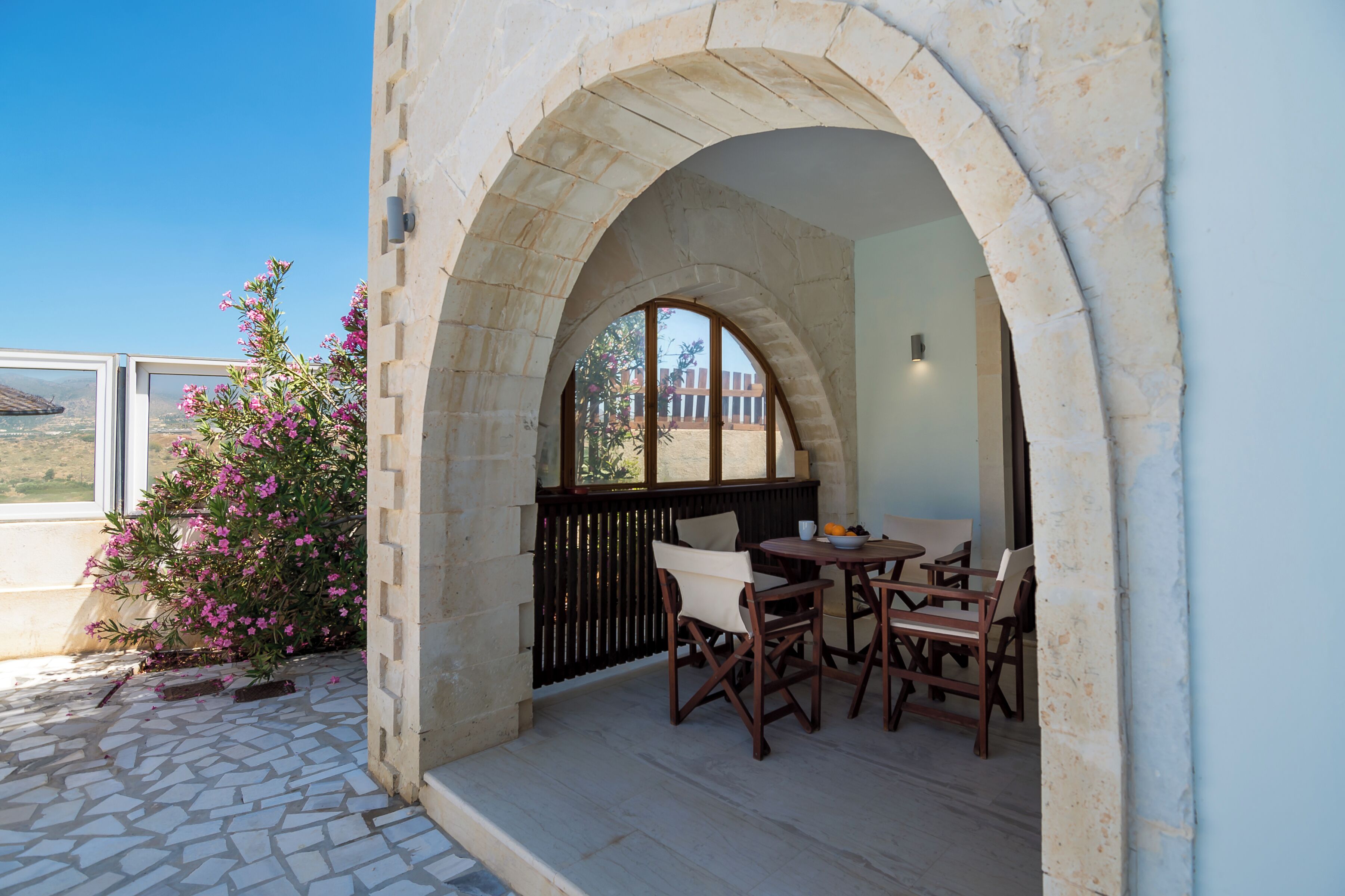 Un patio exterior con suelo de piedra, una mesa y sillas de madera, y una vista a través de una ventana arqueada de piedra.