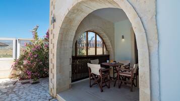 Un patio exterior con suelo de piedra, una mesa y sillas de madera, y una vista a través de una ventana arqueada de piedra.