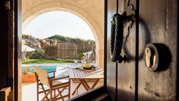 Puerta abierta que da a un patio con piscina, mobiliario de madera y un arco de piedra.