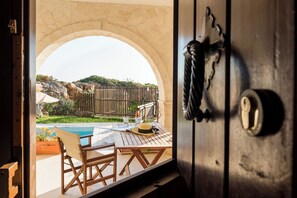 An open door revealing a patio with a pool, wooden furniture, and a stone archway. - Crete Essence Villas – Lagkada (Sitia)