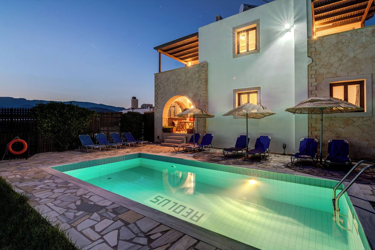 A swimming pool with lounge chairs and umbrellas, a stone-tiled deck, and a building with a stone facade and arched entrance.