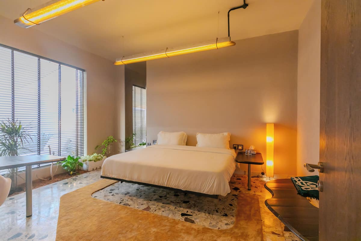 A modern bedroom with a large bed, bedside table, and a nightstand. There is a window with blinds, a potted plant, and a rug on the floor.