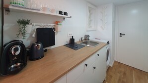 Una cucina compatta con un piano di lavoro in legno, una macchina da caffè, un bollitore e un lavandino. Sui pensili si trovano tazze e una pianta in vaso.