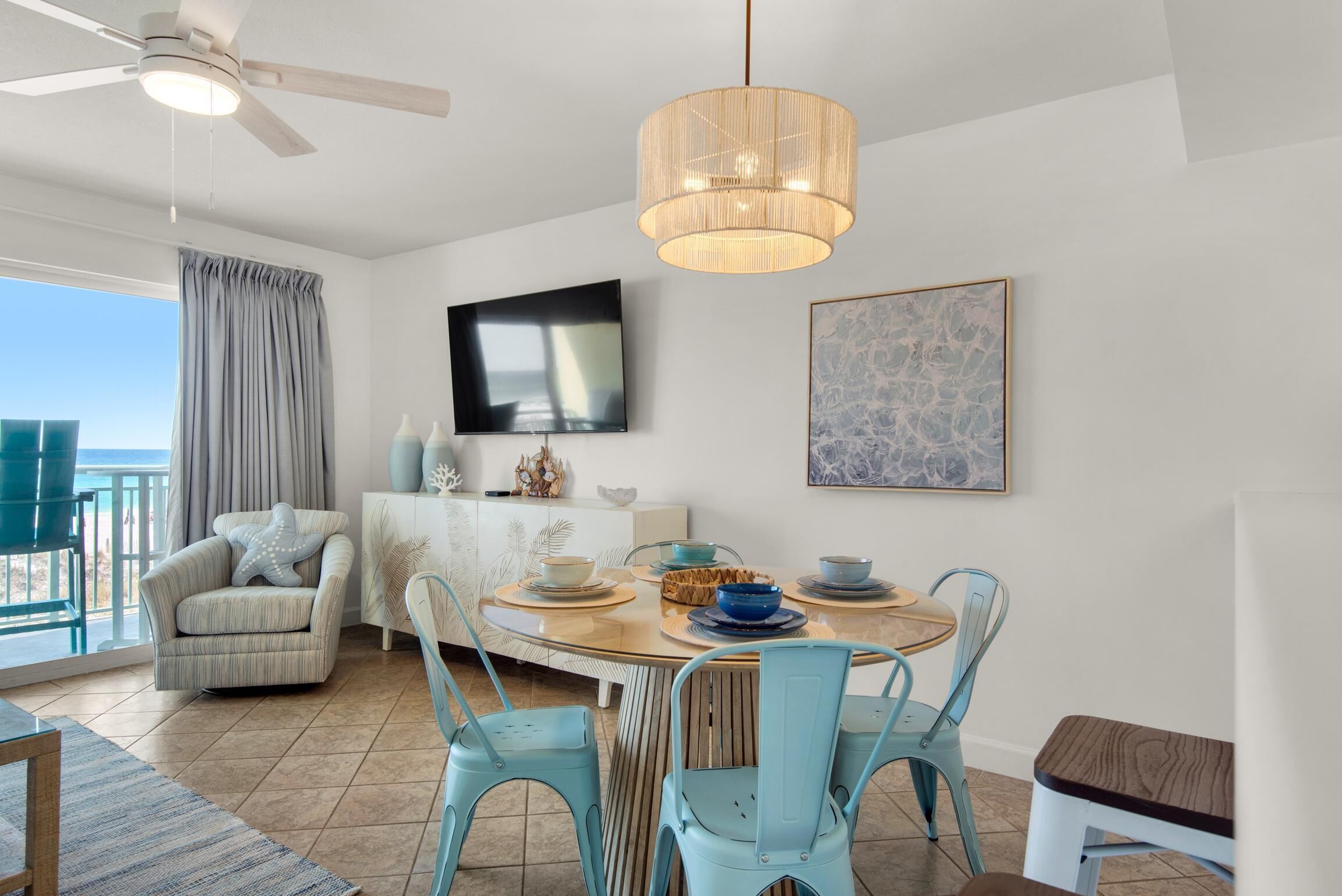 Un coin repas avec une table ronde, des chaises bleues, un ventilateur de plafond et un téléviseur fixé au mur.
