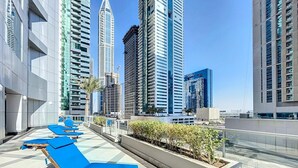 Un espace de détente aménagé sur le toit, avec des chaises longues bleues, un jardinier contenant de petites plantes et une vue panoramique sur la ville, avec ses nombreux immeubles.