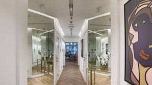 Un couloir aux murs recouverts de miroirs, un parquet en bois à motifs chevrons et une table en verre ornée de fleurs blanches.