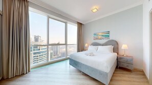 A modern hotel room with a large window offering a city view, a neatly made bed, and a small bedside table with a lamp.