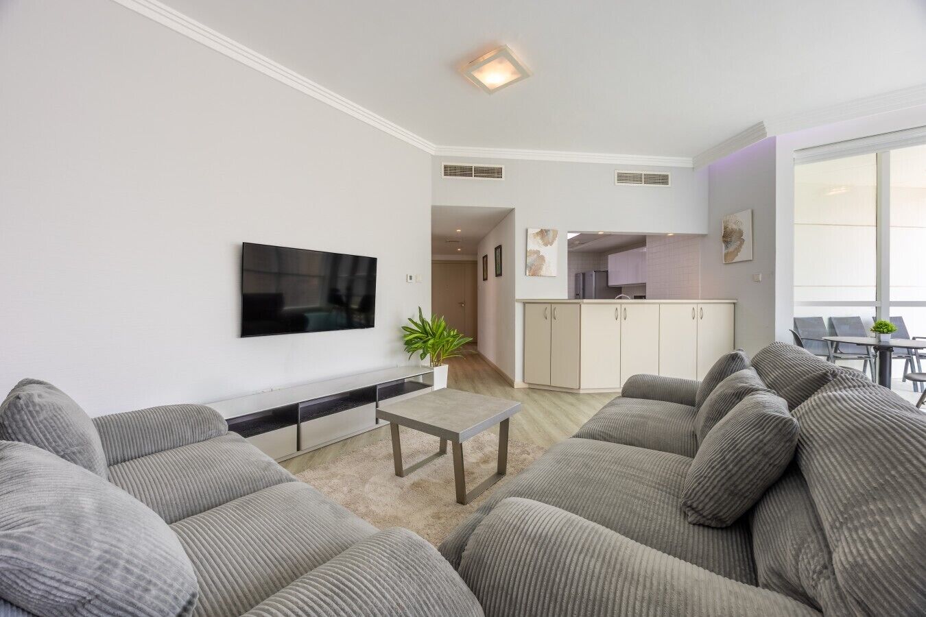 Een moderne woonkamer met een flatscreen televisie, grijze banken, een salontafel en een keuken op de achtergrond.