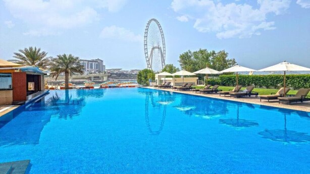 Una piscina con lettini e ombrelloni, una ruota panoramica sullo sfondo e un cielo sereno.
