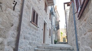 Μια στενή πέτρινη σκάλα οδηγεί σε μια πόρτα σε ένα ιστορικό κτίριο, το οποίο διαθέτει ξύλινα παντζούρια και μεταλλική μπαλκονόπορτα.