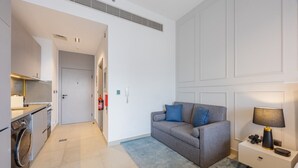 A modern apartment with a kitchenette, a grey sofa with blue cushions, a bedside table with a lamp, and a fire extinguisher on the wall. - Mag 980 - Cosy Studio with City Views in Meydan (Meydan)