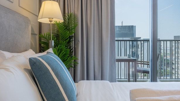 A hotel room with a bed, a lamp, curtains, and a view of the city through large windows. - Mag 980 - Cosy Studio with City Views in Meydan (Meydan)