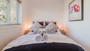 Un lit bien fait, agrémenté de serviettes pliées, deux tables de chevet avec des lampes, une fenêtre avec des stores et une œuvre d’art florale encadrée au mur.