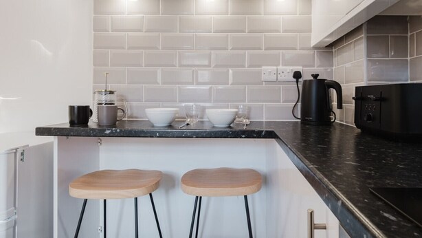 Une cuisine moderne avec des carreaux de céramique blancs disposés en motif métro, un plan de travail noir, deux tabourets en bois et une machine à café.