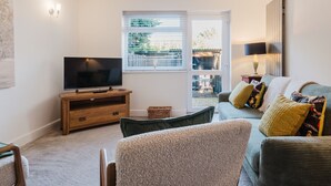 Un salon avec un téléviseur posé sur un meuble en bois, un canapé avec des coussins, une fenêtre avec des stores et un radiateur.
