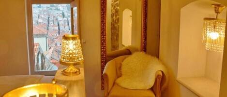 Una habitación con vista a un paisaje urbano a través de una ventana, un candelabro, un espejo y un sillón con un almohadón mullido.