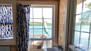 Une cabane avec un intérieur en bois, une fenêtre donnant sur un lac, un lavabo et une chaise près de la fenêtre.