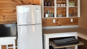 Une cuisine compacte avec un réfrigérateur blanc, un four à micro-ondes et un petit plan de travail sur lequel se trouve un grille-pain. Au-dessus du plan de travail, on trouve un meuble en bois contenant divers ustensiles de cuisine.