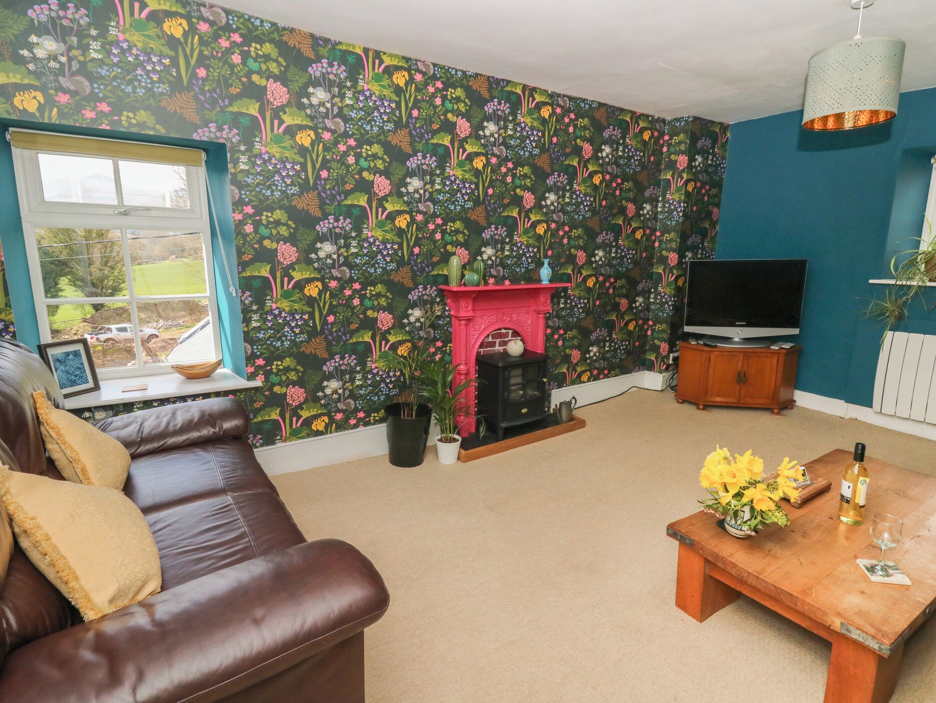 A living room with a floral wallpaper, a brown leather sofa, a wooden coffee table with flowers, a fireplace, and a television.