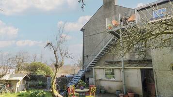 A house with a visible staircase leading to a second floor, a small garden with chairs and potted plants, and a clear sky.
