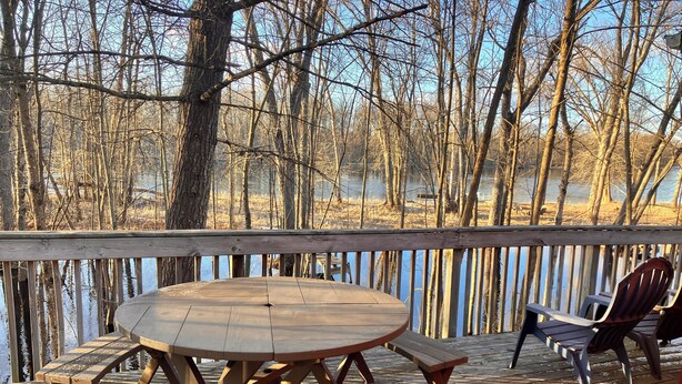 A wooden deck with a round table and benches, overlooking a body of water and surrounded by trees.