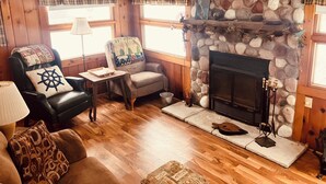 A cozy living room with a stone fireplace, wooden walls, and a comfortable seating area with a leather sofa and armchairs.