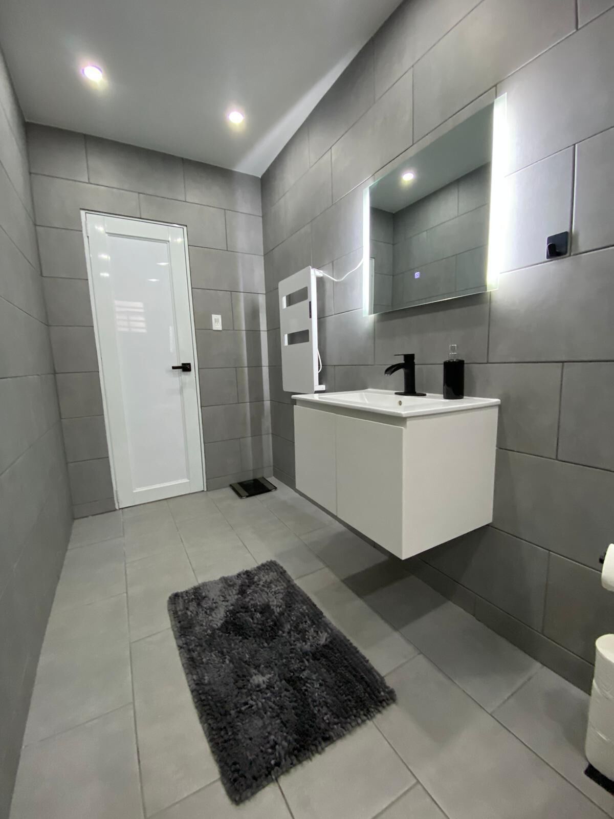 Een moderne badkamer met grijze tegels, een wit badkamermeubel, een zwarte kraan, een spiegel en een grijze vloerkleed.