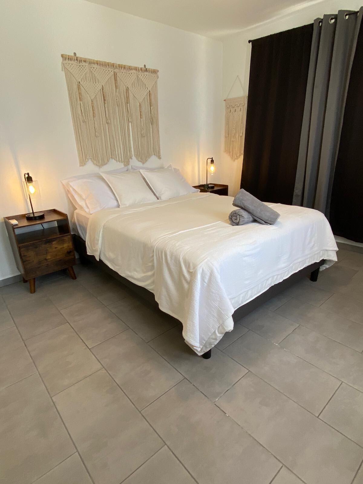 A neatly arranged bedroom with a bed, two bedside tables, and a headboard with macrame curtains.