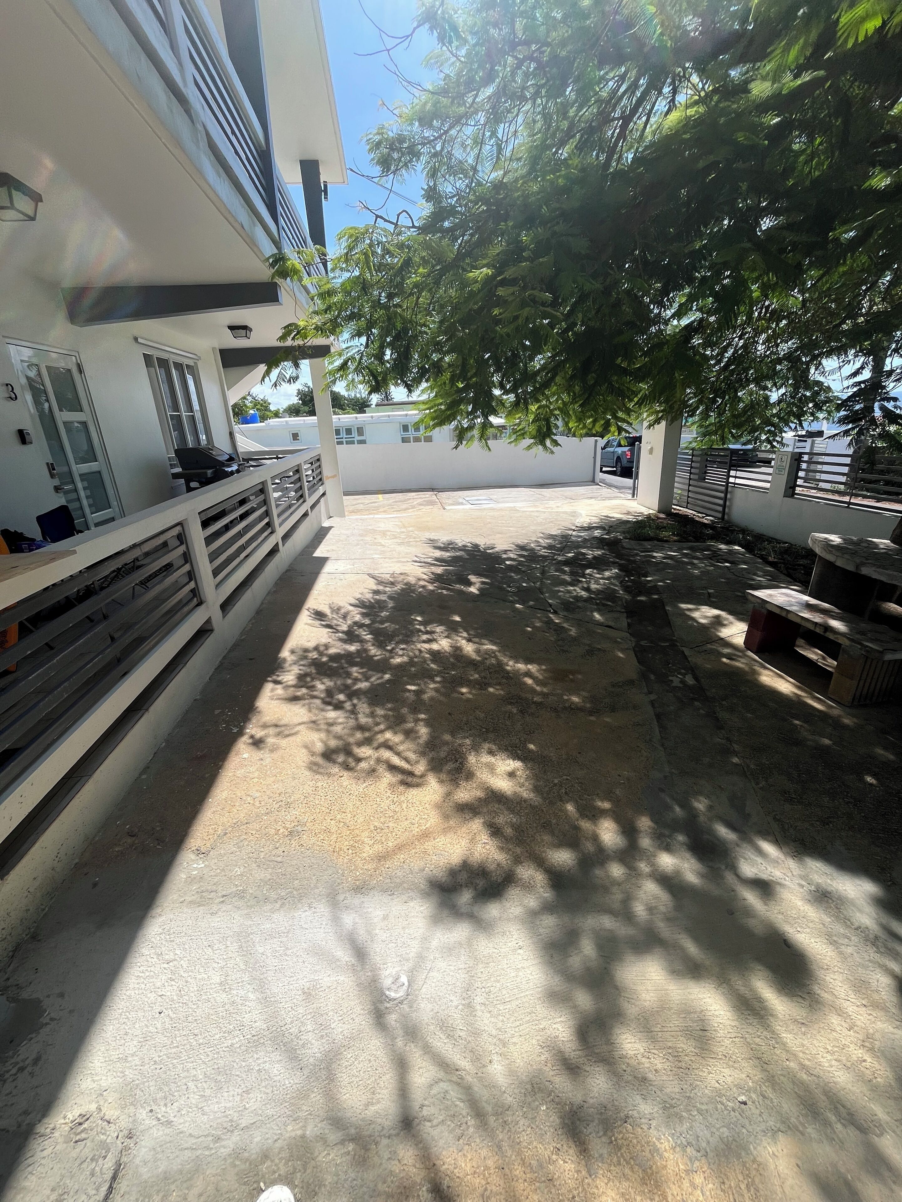 A covered outdoor area with a concrete floor, a white railing, and a tree providing shade.
