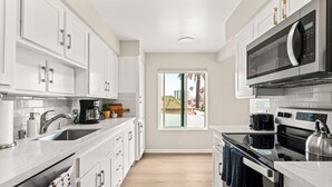 A modern kitchen with white cabinets, stainless steel appliances, and a view of palm trees through a window. - Seize the Day by Gather: 2BR Retreat w/ Views & Parking (La Jolla)