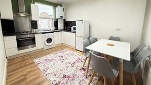 A compact kitchen with a washing machine, a microwave, and a refrigerator. There is a dining table with chairs and a rug on the floor.