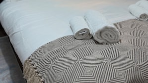 A neatly made bed with white linens and rolled towels, a patterned throw blanket, and a framed picture on the wall.