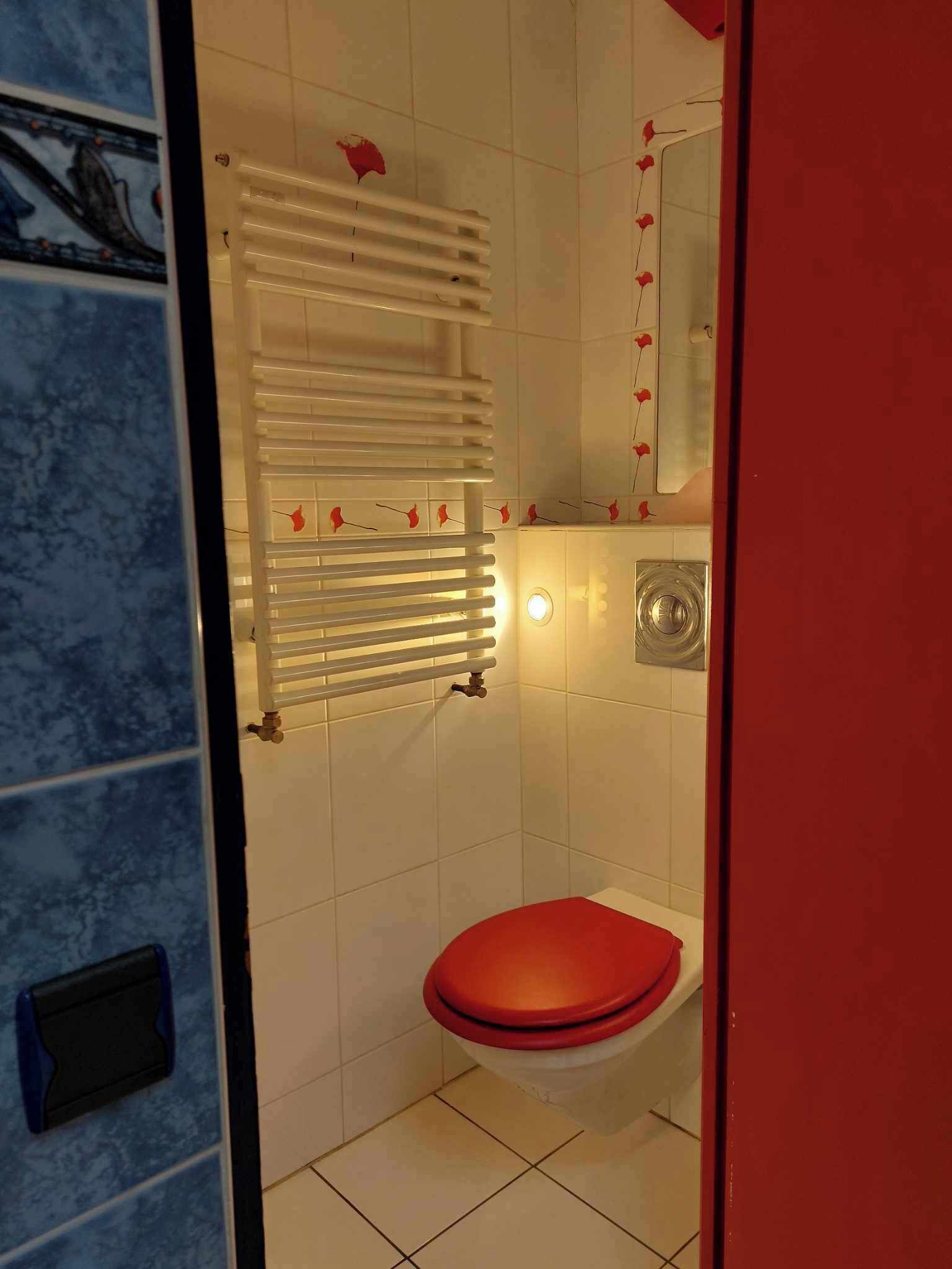 A bathroom with a red toilet, white tiled walls, a white towel rack with red flowers, and a round mirror.