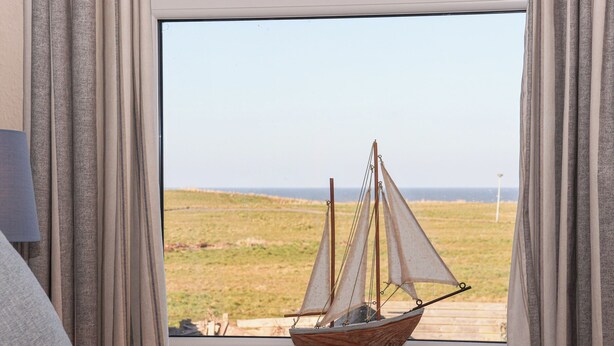 A model sailboat on a windowsill with a view of a grassy field and the sea. - 48 Waxholme Road (Withernsea)