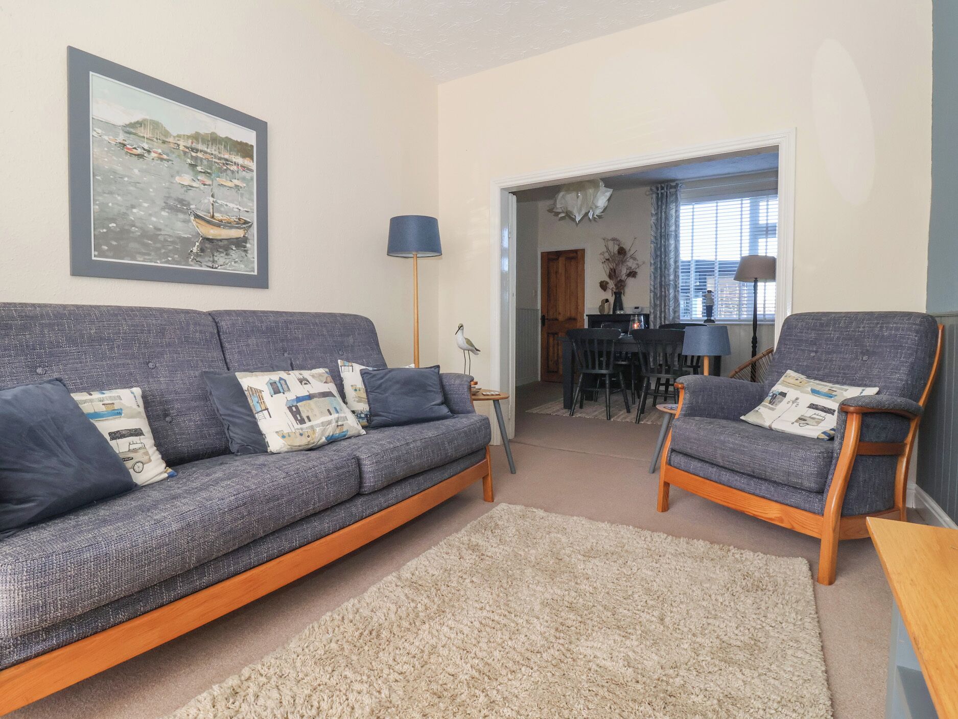 Ein Wohnzimmer mit grauem Sofa, zwei Sesseln, einem beigen Teppich, einem Esstisch mit Stühlen und einem gerahmten Bild an der Wand.