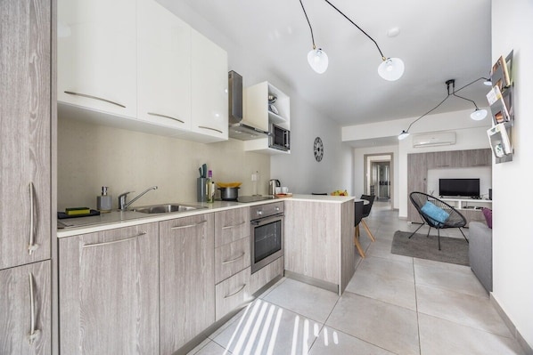 A modern kitchen with light wood cabinets, a built-in oven, and a large island with a sink. There is a dining area with a chair and a TV in the background. - Modern Downtown Apartment (Gżira)