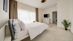 Quarto de hotel moderno com cama grande, televisão de tela plana, mesa de cabeceira, planta em vaso e vista para o corredor.