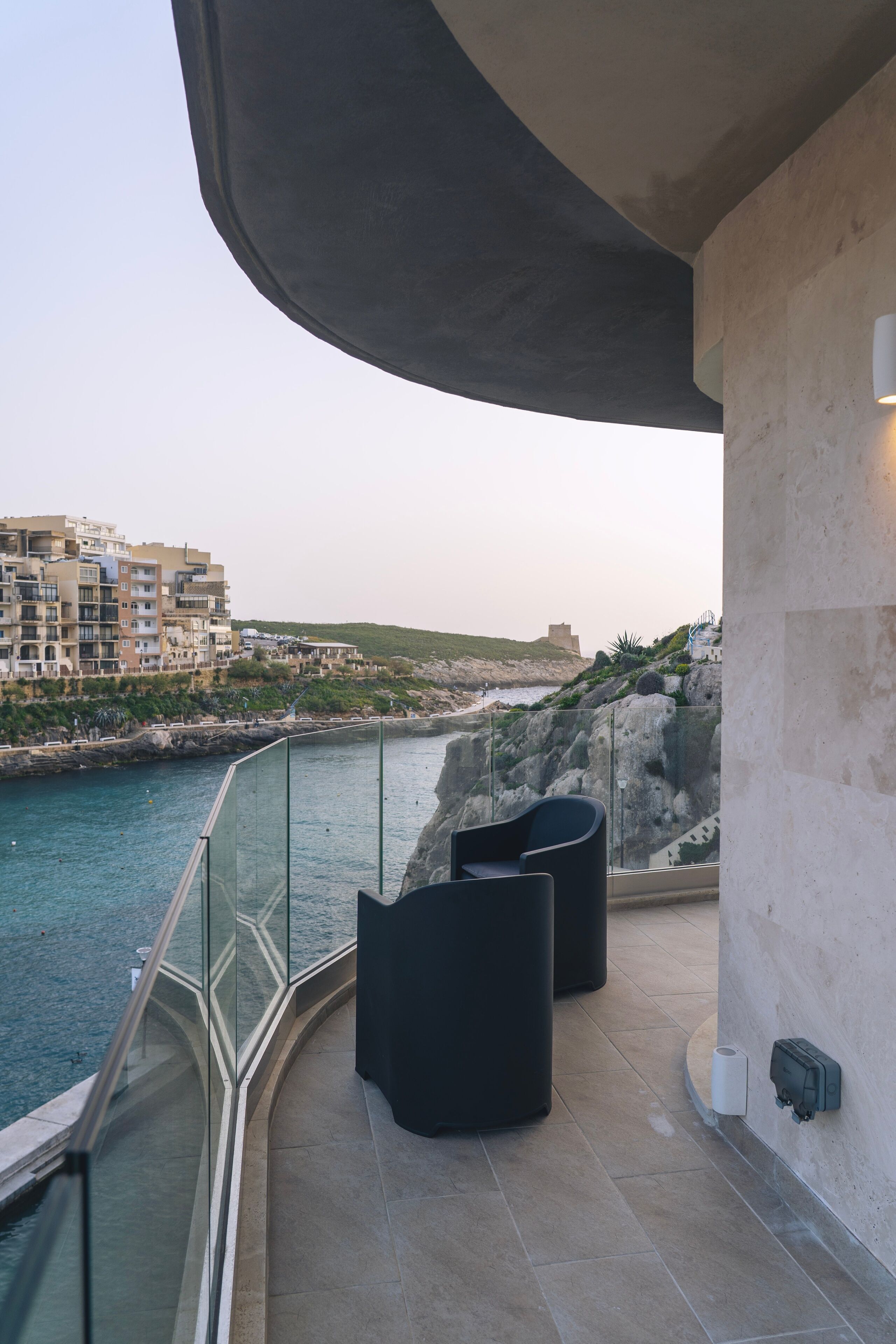 Un balcon doté d’une balustrade en verre, donnant sur une zone côtière, avec un espace salon comprenant des chaises noires et une vue sur les bâtiments et la mer.