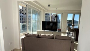 A modern hotel room with a large bed, a sofa, a TV, and a dining area with a view of the city.