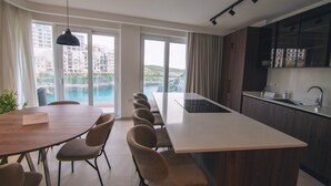 A modern kitchen with a central island, a dining area with a long table and chairs, and large windows offering a view of buildings and water.