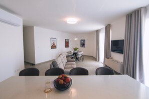 A modern living room with a sofa, dining table, and a TV. There are framed pictures on the wall, a ceiling light, and a window with curtains.
