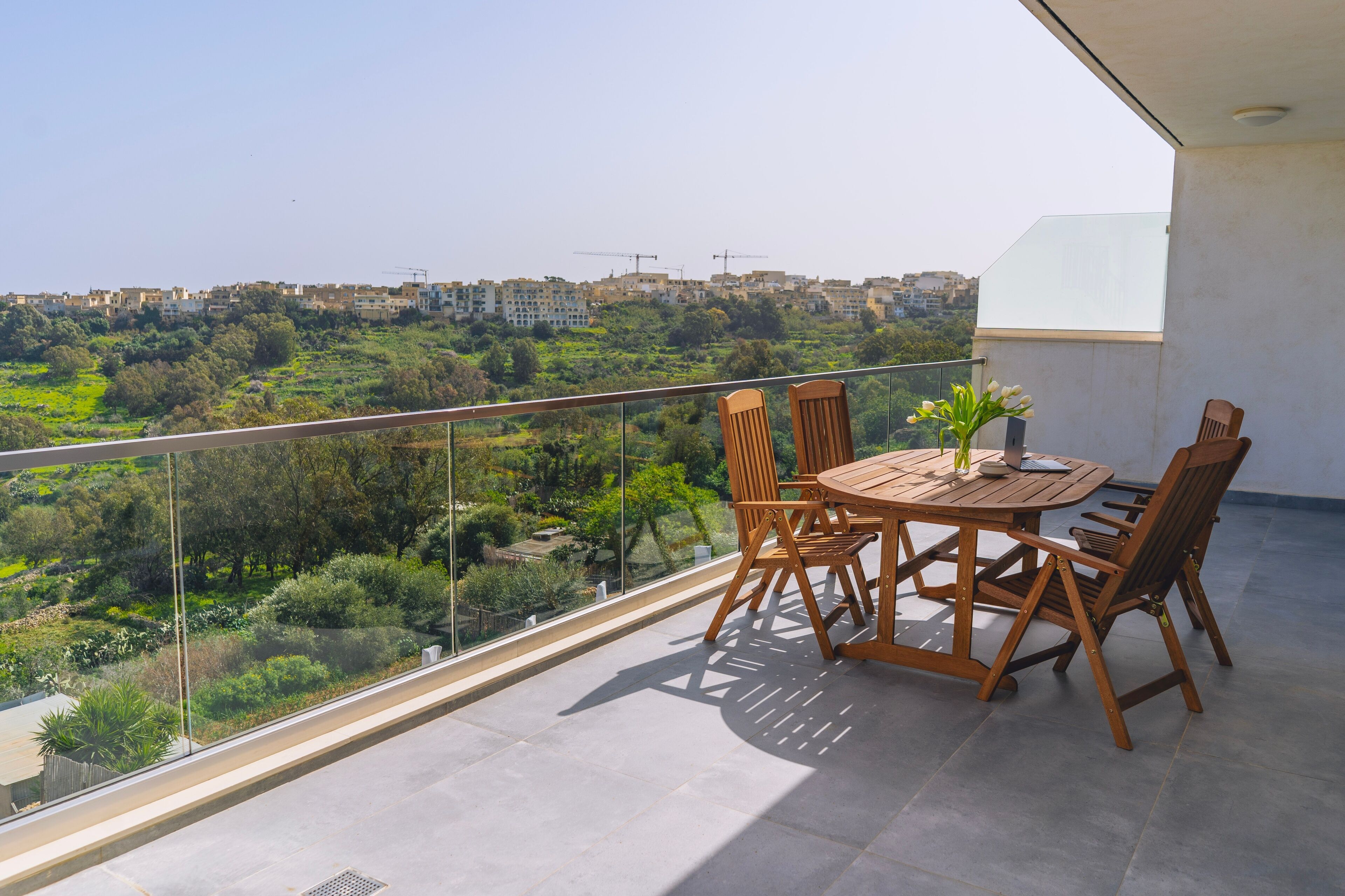 Un balcone con tavolo e sedie, affacciato su una città e aree verdi.