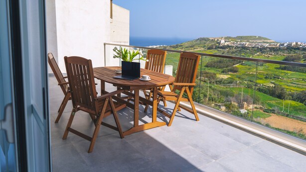 Een balkon met een houten tafel en stoelen, een bloempot en uitzicht op een kustlandschap.
