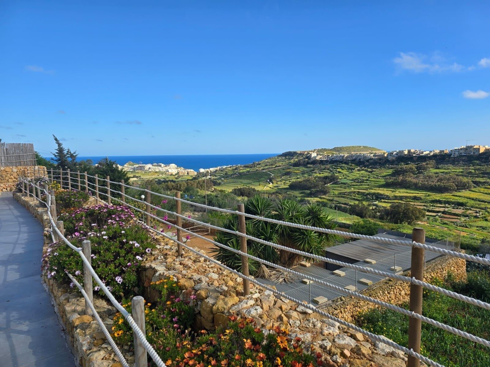 A scenic coastal view with a stone pathway, lush greenery, and a clear blue sky.