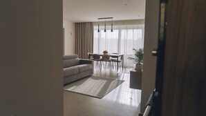 Un salon moderne avec un canapé, une table à manger et des chaises. Une grande fenêtre, ornée de rideaux, laisse entrer la lumière, et une plante est placée sur le côté.