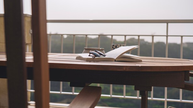 Une table en bois sur laquelle repose un livre ouvert et une paire de lunettes, le tout placé devant la rambarde d’un balcon d’où l’on peut admirer la verdure.